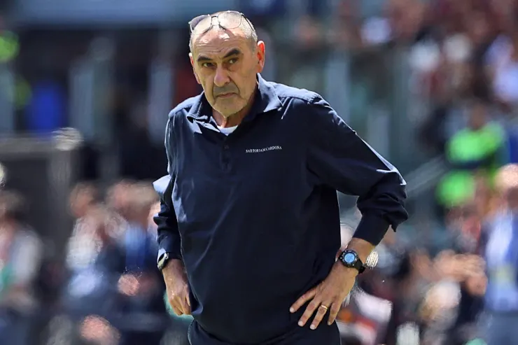 Maurizio Sarri in piedi a bordo campo durante una partita, con occhiali sulla testa e abbigliamento sportivo blu scuro