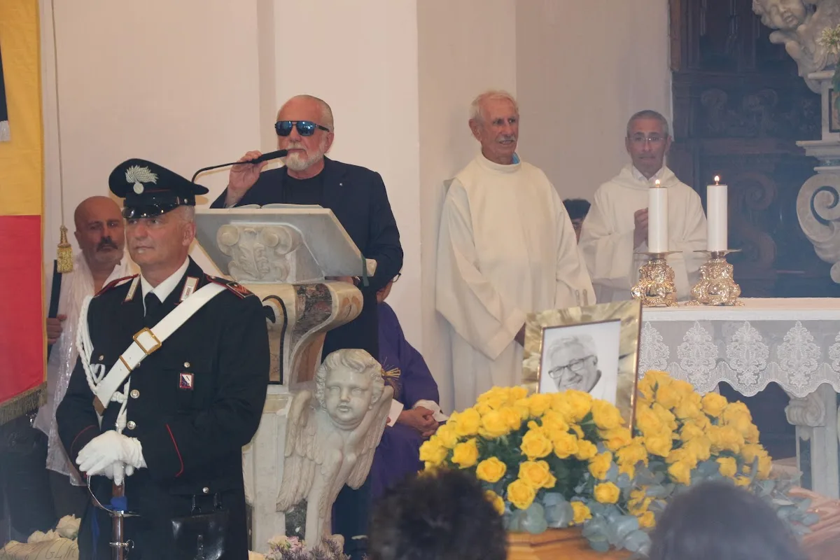 Aurelio De Laurentiis in abito scuro durante una cerimonia religiosa con carabinieri e sacerdoti, fiori gialli in primo piano
