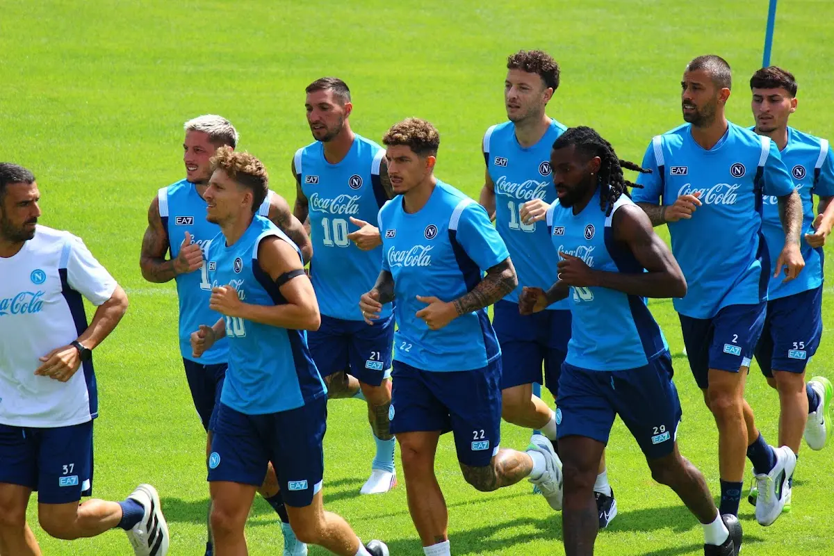 Giocatori del Napoli in allenamento corrono insieme sul campo in divisa blu con sponsor Coca-Cola