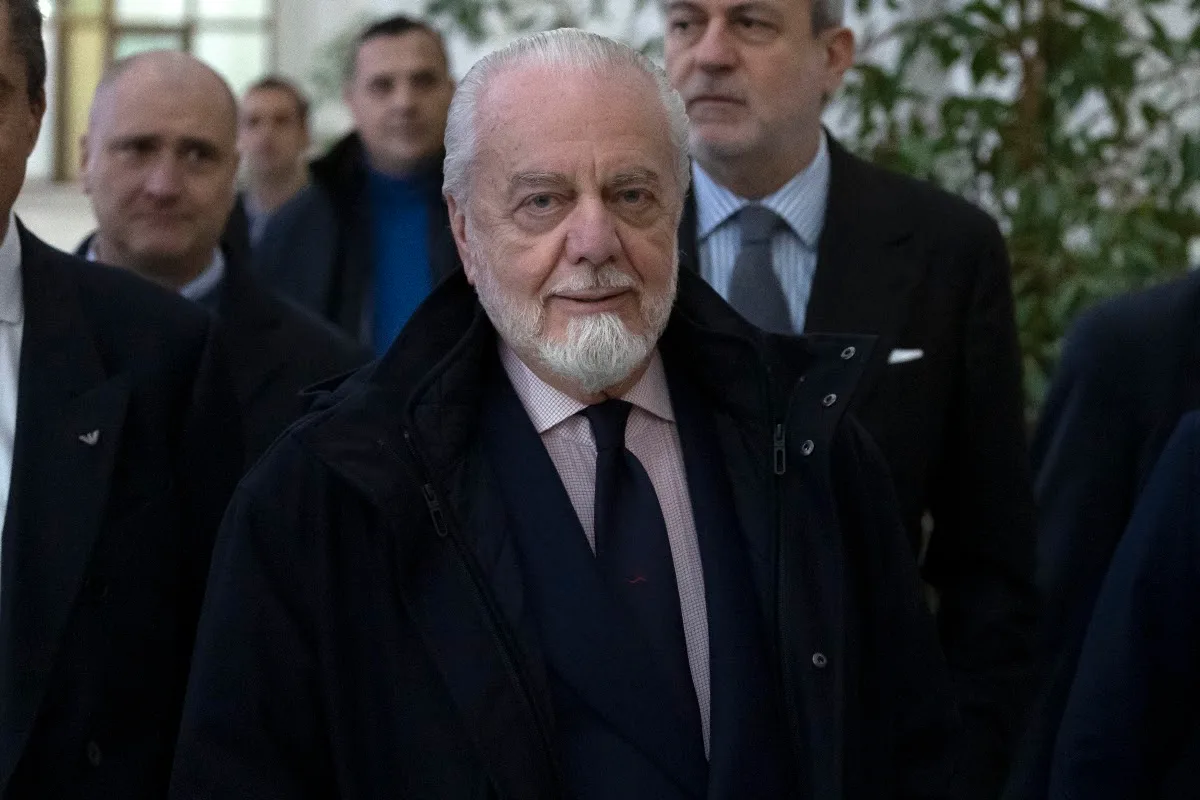 Aurelio De Laurentiis con la barba, in abito scuro e cravatta, circondato da collaboratori in ambiente interno