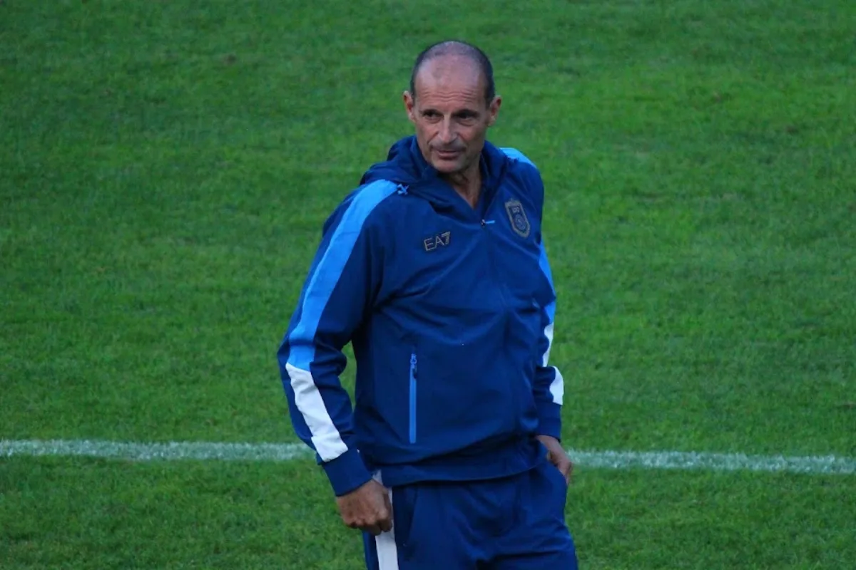 Uomo calvo in tuta blu con dettagli azzurri e logo EA7 su campo da calcio con prato verde