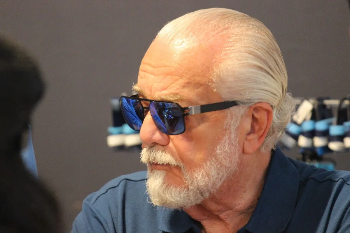 Uomo con capelli e barba bianchi, occhiali da sole con lenti a specchio blu e polo blu, in primo piano