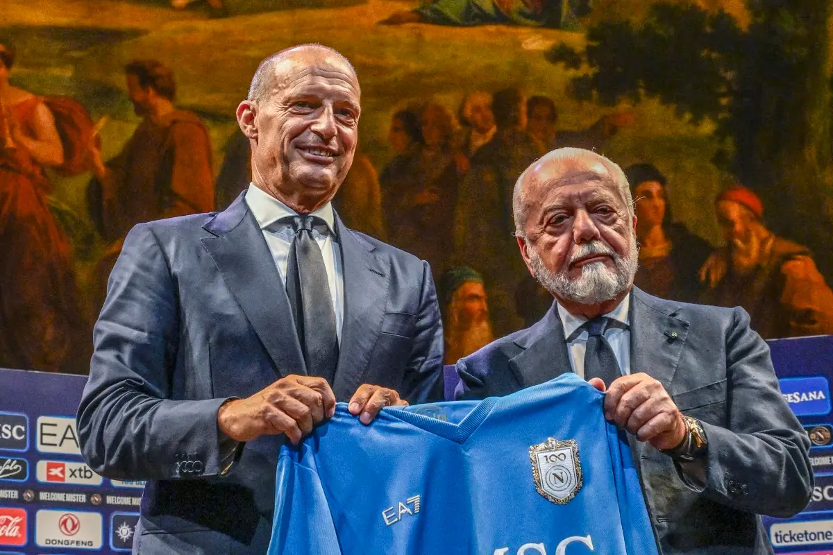 Due uomini in giacca scura tengono una maglia azzurra del Napoli con lo stemma del centenario davanti a un fondale con dipint