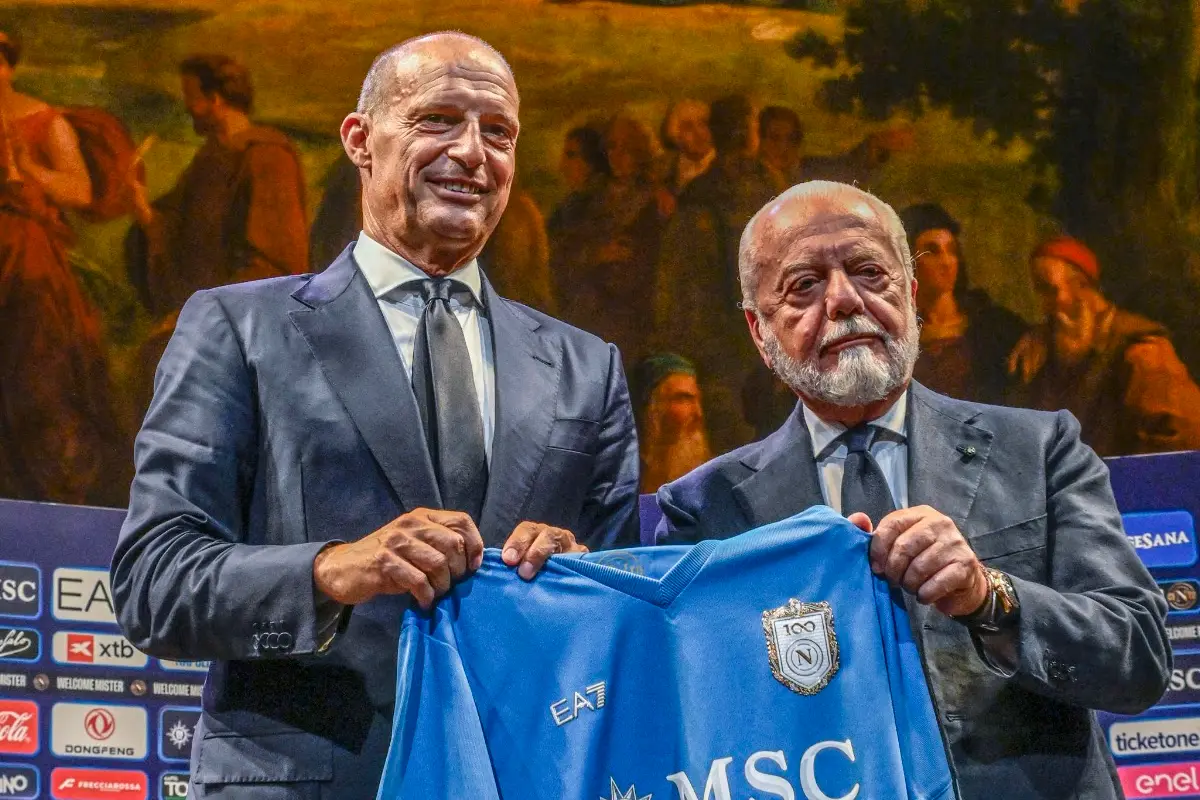 Due uomini in giacca scura reggono una maglia azzurra del Napoli con lo stemma del centenario e lo sponsor MSC davanti a un p