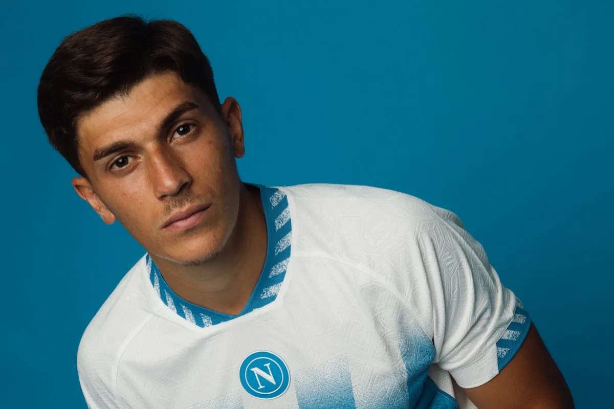 Giocatore del Napoli in maglia bianca e blu con logo SSC Napoli su sfondo azzurro
