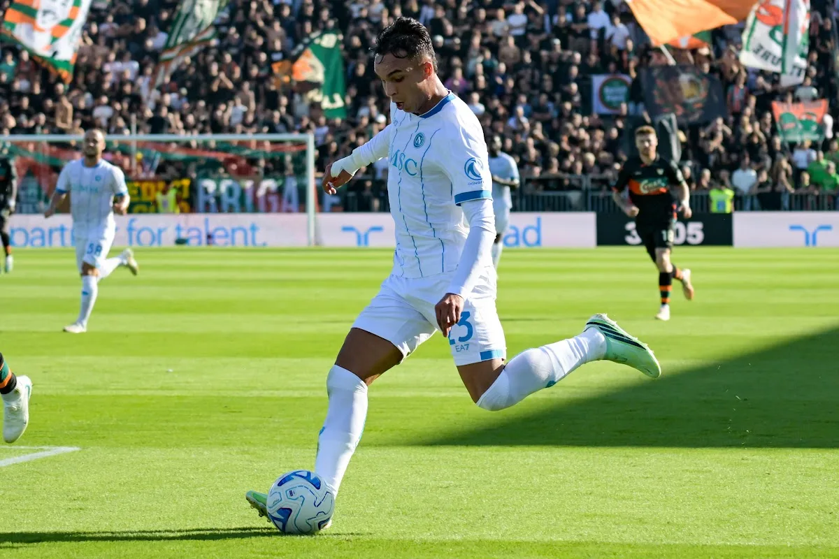 Giovane del Napoli in maglia bianca numero 23 calcia il pallone durante una partita, con tifosi sugli spalti