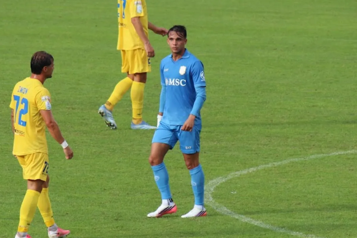 Giovane con la maglia azzurra del Napoli in campo durante una partita, accanto a un avversario in giallo
