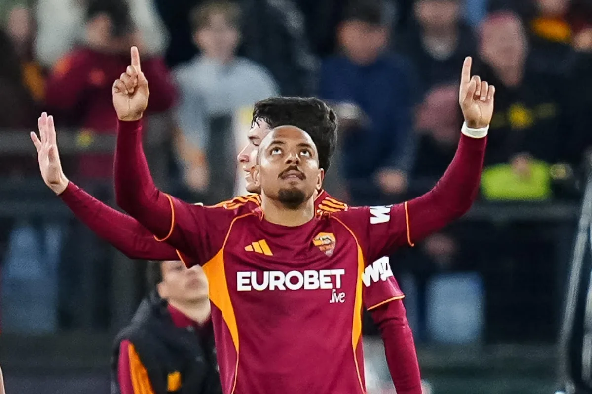 Giocatore della Roma con maglia rossa e gialla esulta alzando le braccia e indicando il cielo durante una partita